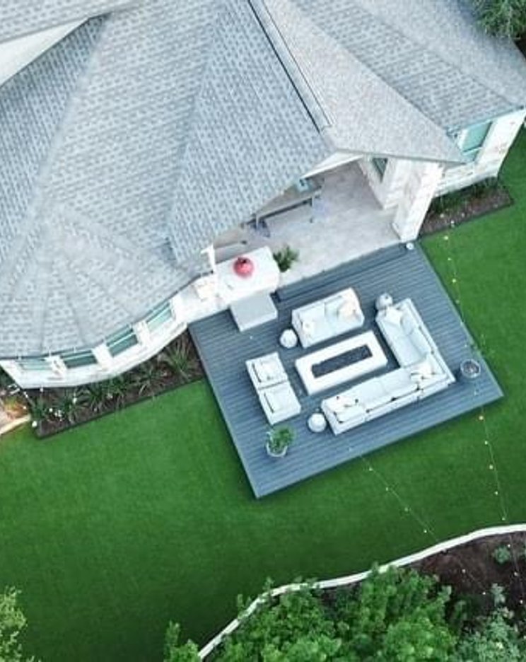 Aerial view of a Bee Cave backyard with a green turf island, deck, and flagstone patio under live oaks