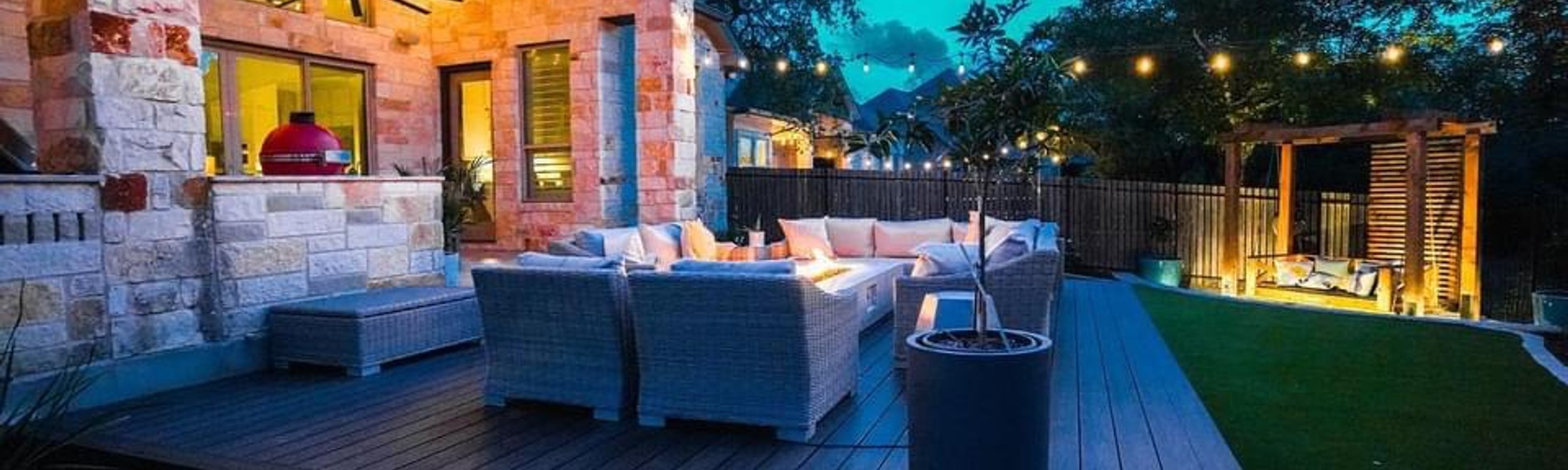 Bee Cave backyard at night with string lights over a deck, fire pit, and turf under the oaks