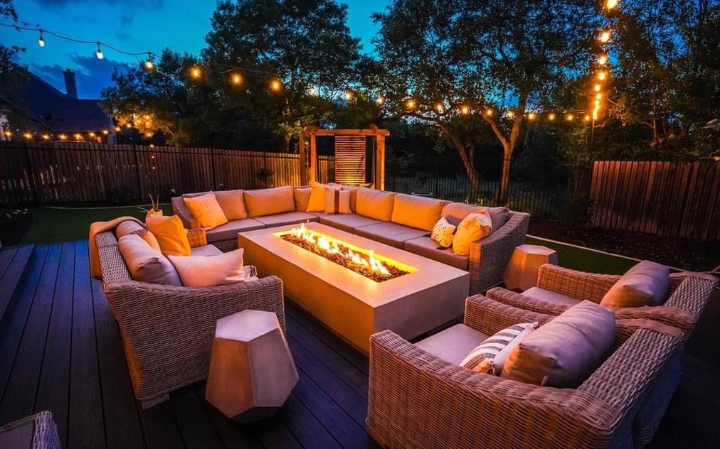 Bee Cave backyard at night with a lit fire pit, string lights over the deck, and cedar privacy screens washed with low voltage landscape lighting