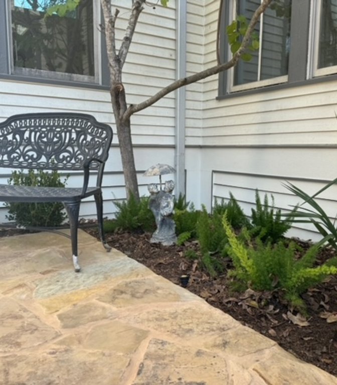 Mulched garden bed beside a flagstone patio and iron bench in a Central Texas garden