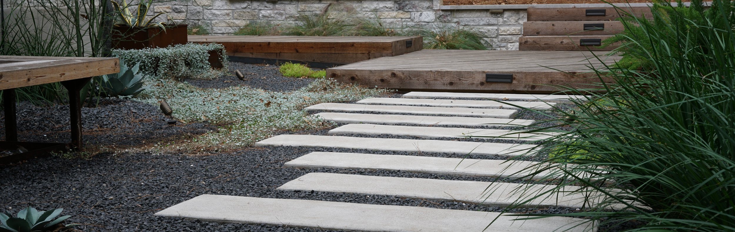Bastrop, TX Lost Pines courtyard with paver walkway, stone retaining wall, and deck by Thrive Landscape and Design