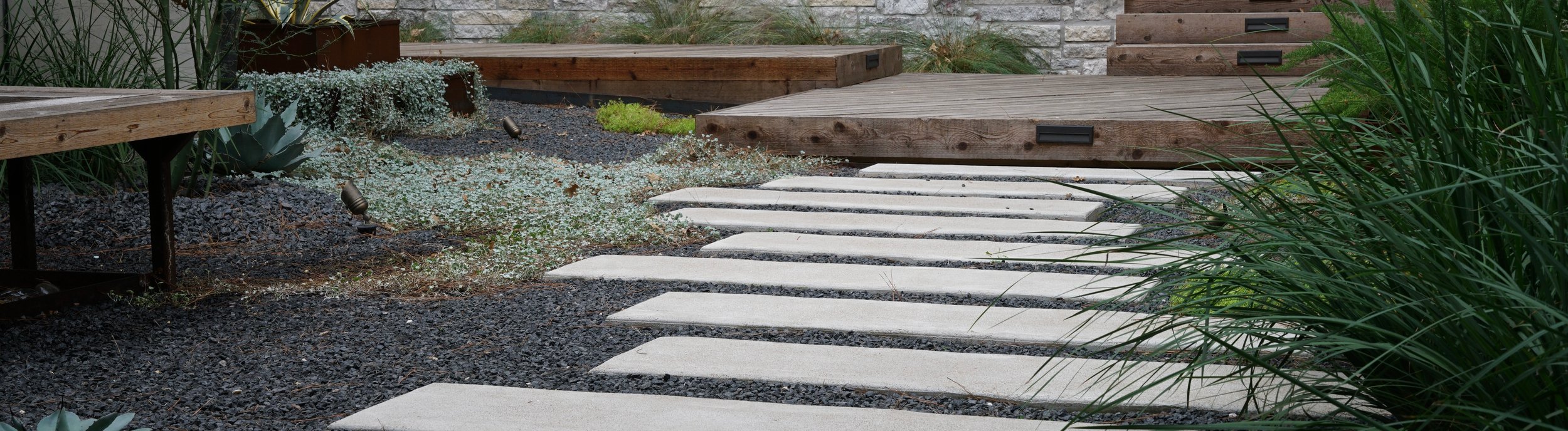 Bastrop, TX courtyard with cream paver walkway, limestone retaining walls, and a deck at the edge of the Lost-Pines forest by Thrive Landscape and Design