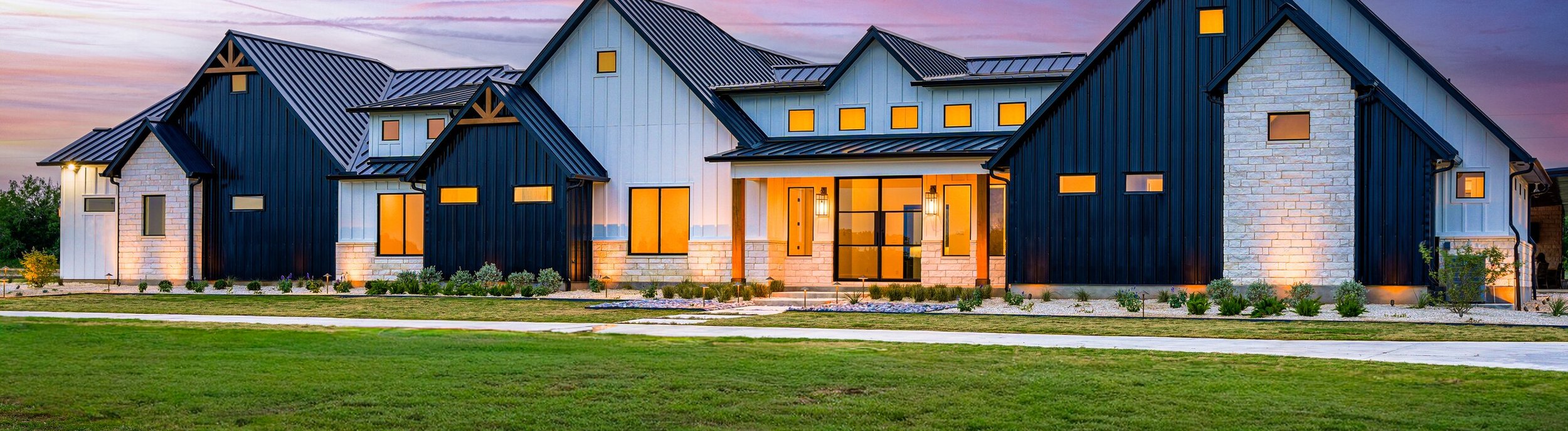 Modern farmhouse front yard at sunset with uplit cream limestone, low path lights, and native plantings in white gravel foundation beds set back from the slab on a new build in Rosanky, Texas