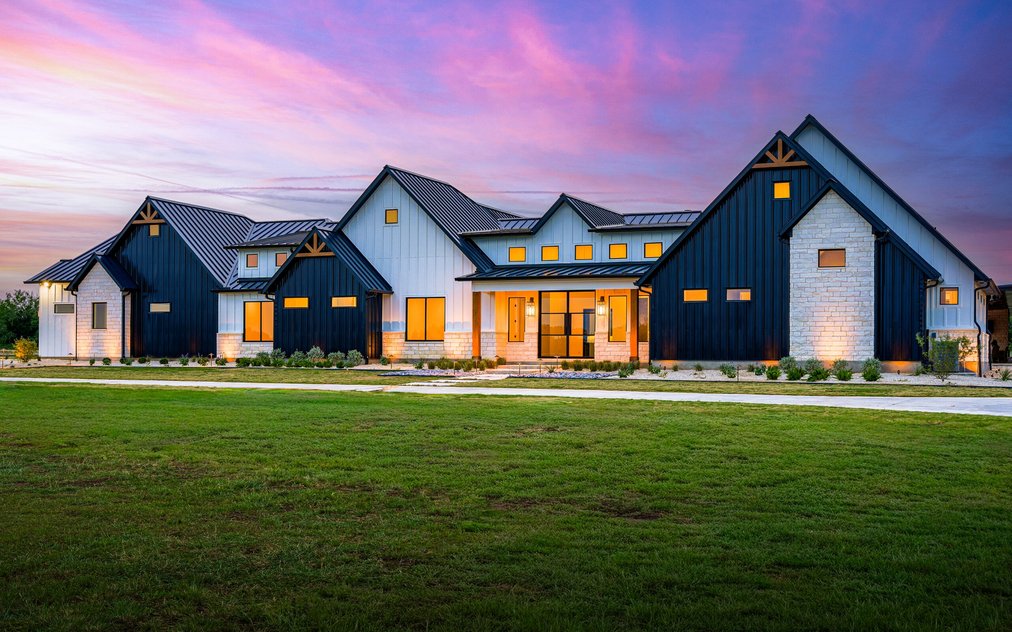 Modern farmhouse front yard at sunset with uplit cream limestone, low path lights, and native plantings in white gravel foundation beds set back from the slab on a new build in Rosanky, Texas