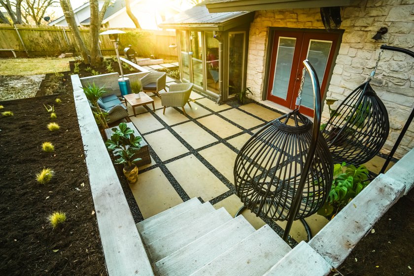 Cream large-format paver patio with black gravel joints, lounge seating, and hanging egg chairs below board-formed concrete retaining walls and concrete steps in a Shoal Creek backyard in central Austin