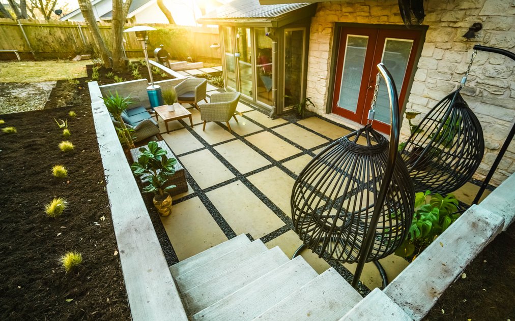 Cream large-format paver patio with black gravel joints, lounge seating, and hanging egg chairs below board-formed concrete retaining walls and concrete steps in a Shoal Creek backyard in central Austin