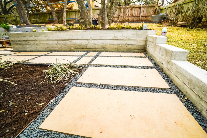 Board-formed concrete retaining wall with built-in steps holding back a raised lawn above a large-format paver and gravel patio in an Austin backyard near Shoal Creek