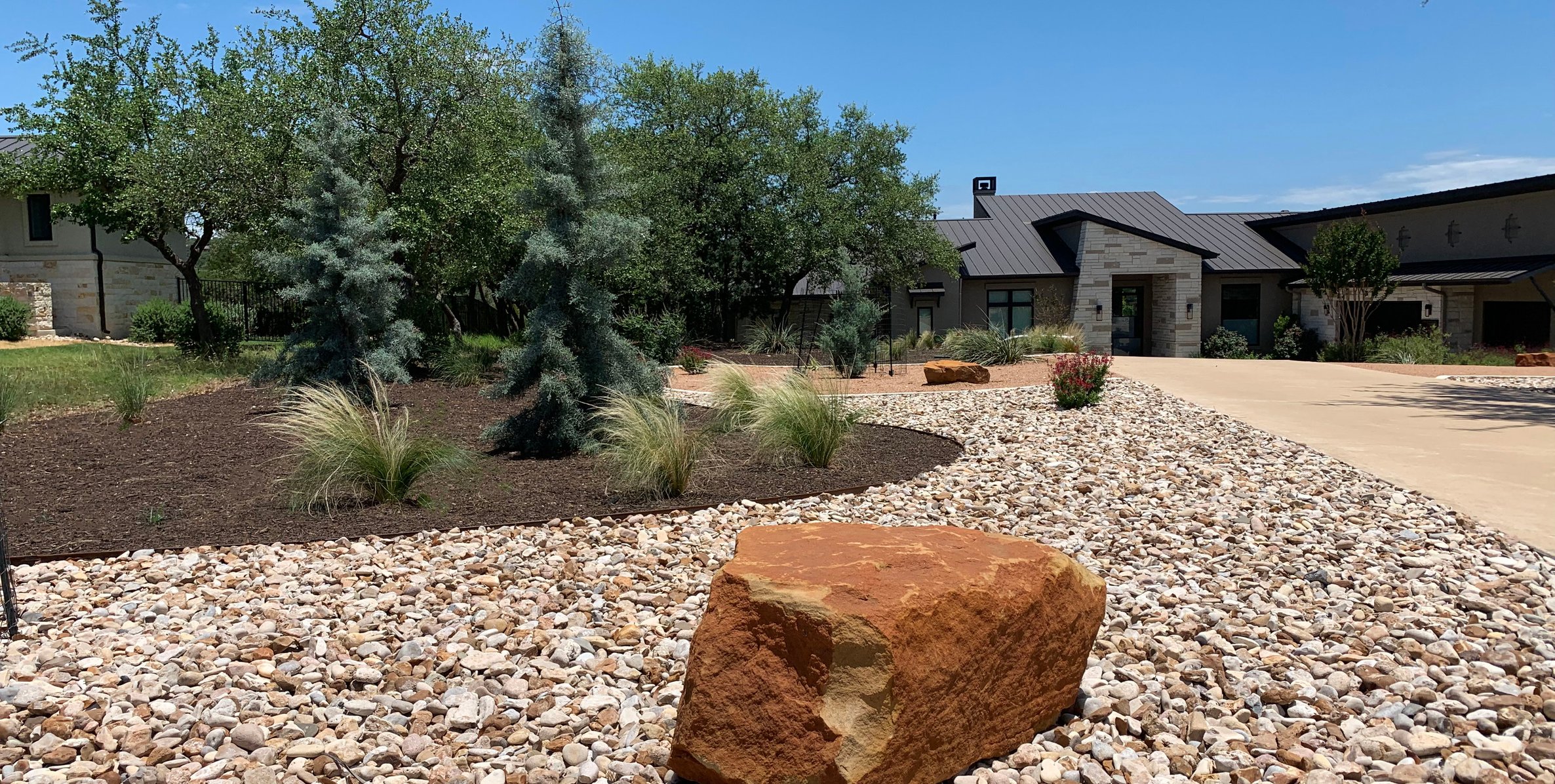 Ground-level view of the river rock zone with a sandstone boulder anchor and blue Arizona cypress accents in front of the home